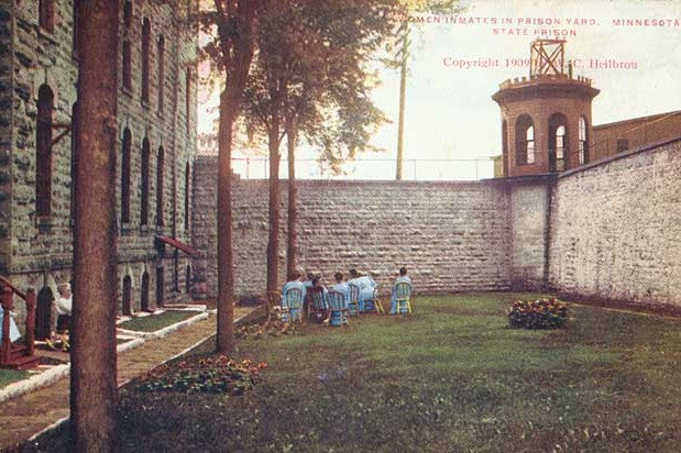 Photo of women in blue sitting in a courtyard below walls and a watch tower. The text reads "Women inmates in prison yard. Minnesota State Prison. Copyright 1909. C. Heilbrou. Photo locator MW4.9 ST7.2S r8 in MNHS Collections