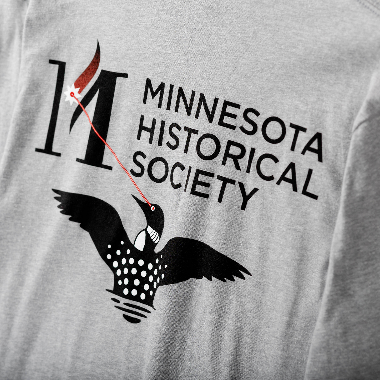 The Long Sleeve T-Shirt Laser Loon is a gray Minnesota shirt with the MNHS logo: a bold black "M," a stylized laser loon, and a red stripe linking the loon's eye to the letter.
