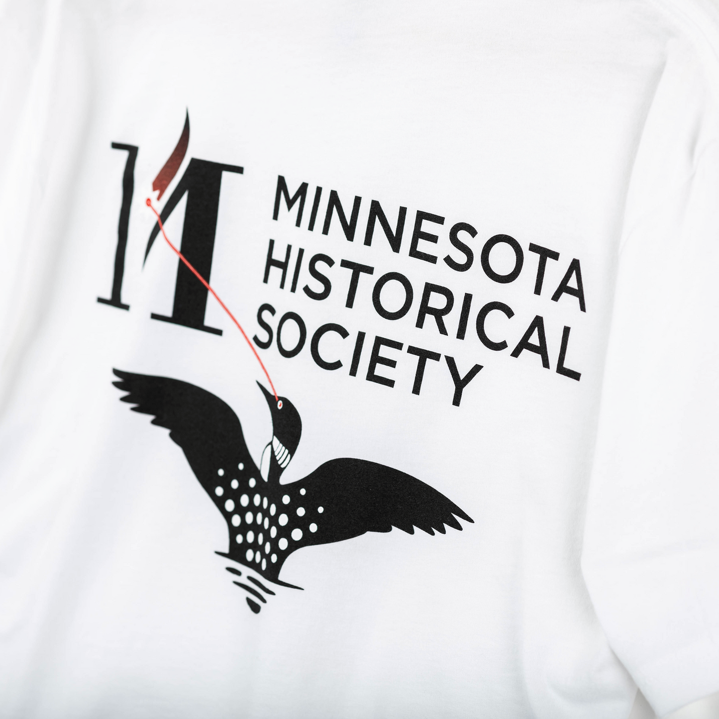 T-Shirt Laser Loon: White Minnesota tee with the MNHS Logo, bold text, and an illustrated black loon with white spots and a red ribbon, flying beneath the organization’s name.