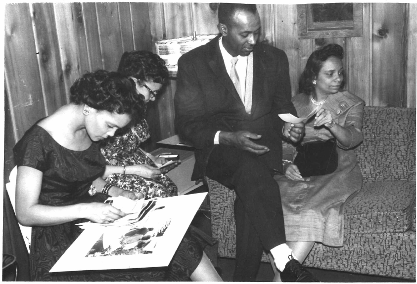 4 people look at photographs; three women and one man. The photo is dated 1955 and is in black and white.