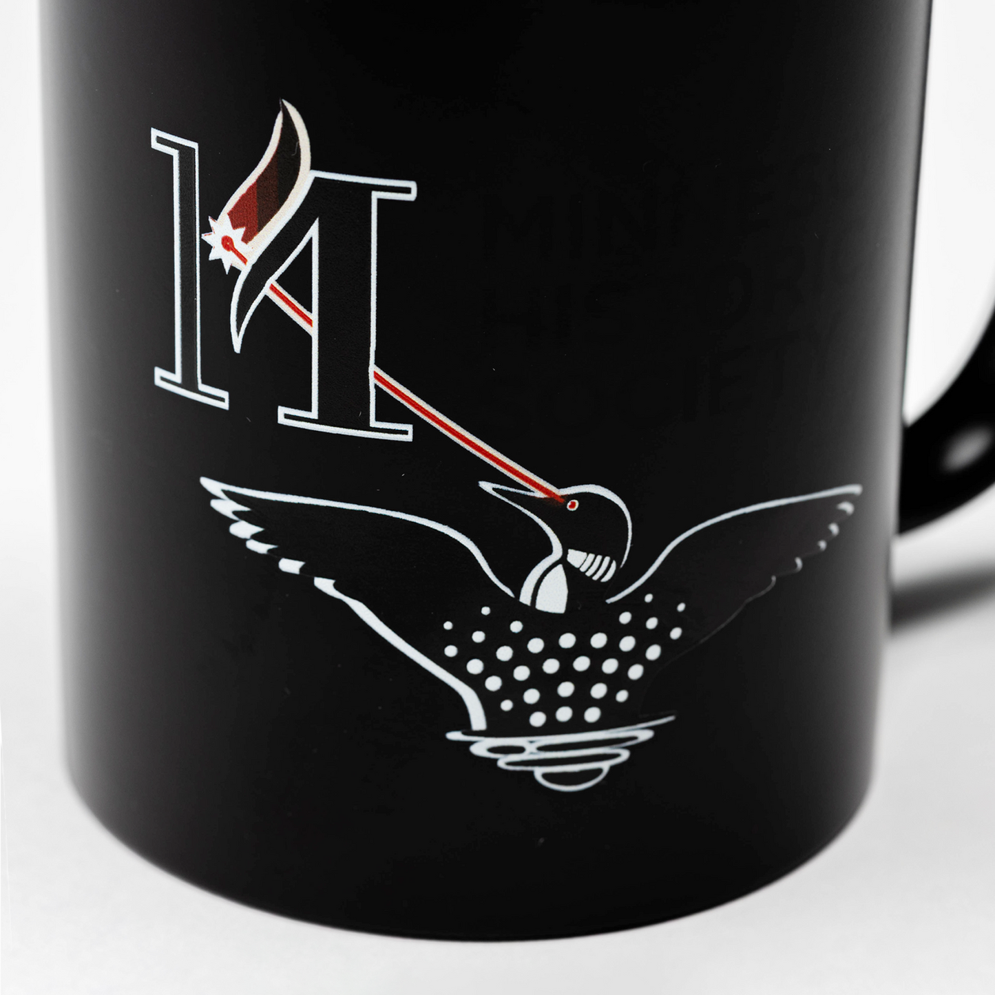 Laser Loon Mug Color Changing features a black mug that reveals a white illustration of the Laser Loon and a bold “M” when hot. The loon’s eye projects a red beam that hits a maple leaf on the “M.” Partial text, reading MINIS and HISTORIC SOCIETY, references the Minnesota Historical Society.