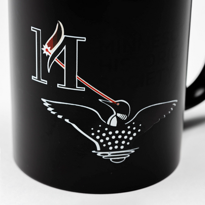 Laser Loon Mug Color Changing features a black mug that reveals a white illustration of the Laser Loon and a bold “M” when hot. The loon’s eye projects a red beam that hits a maple leaf on the “M.” Partial text, reading MINIS and HISTORIC SOCIETY, references the Minnesota Historical Society.