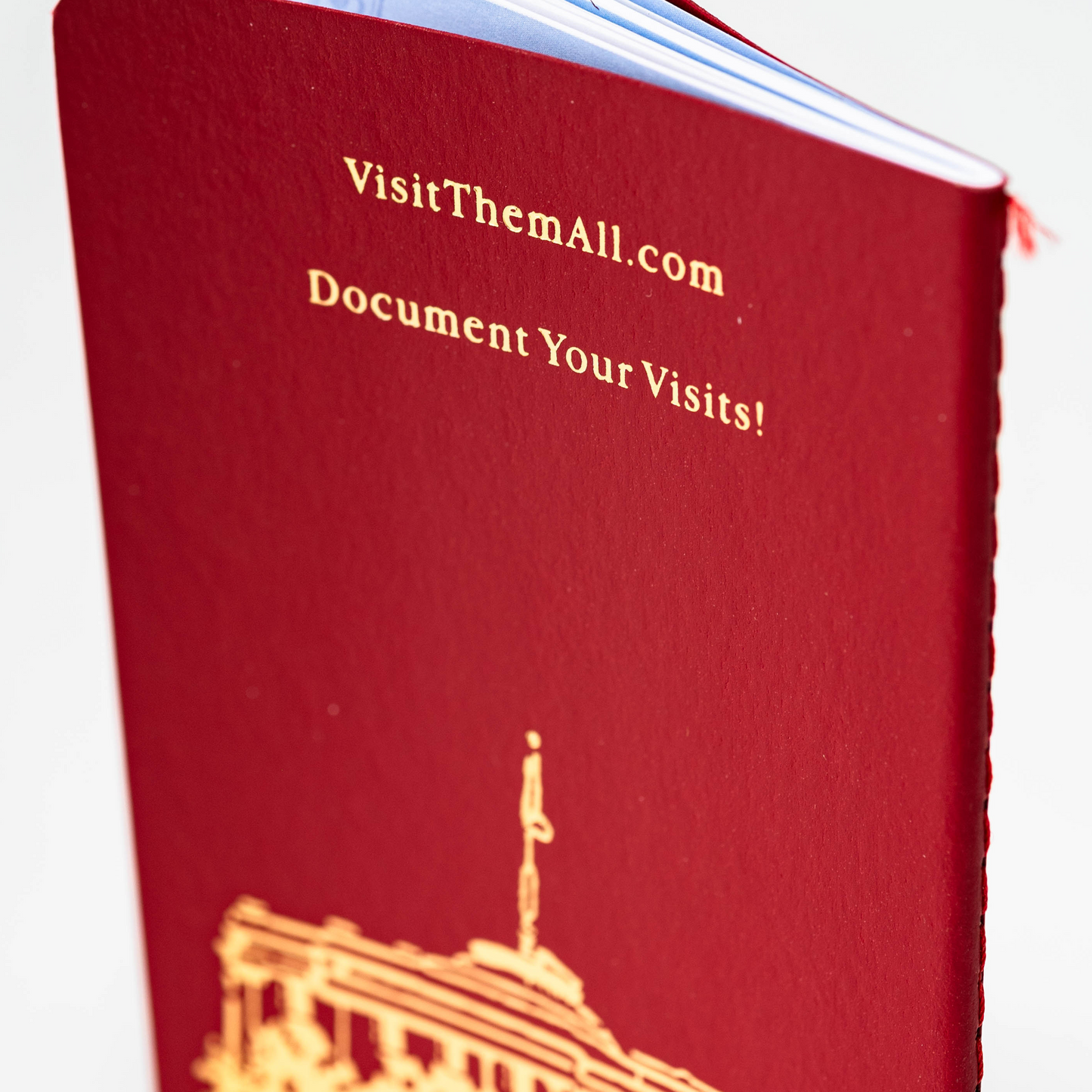 A close-up of the State Capitol Passport—a red booklet with gold text reading VisitThemAll.com Document Your Visits! and part of a gold illustration of a building on the cover. The blue-tinted edge is visible, highlighting its passport-like appearance.
