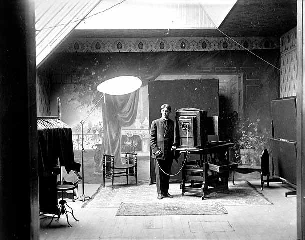Title: Photographer in the Paul Engstrom studio at Cannon Falls. 1910. Paul Engstrom. MNHS locator: N2.1 r13. Description:  A photographer stands with an early 1900s camera. He's holding the wired control for the camera. Behind him is a painted backdrop of a garden or fancy sitting room.