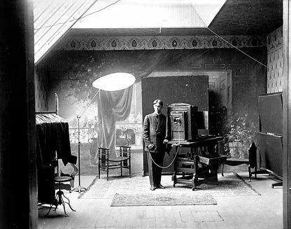 Title: Photographer in the Paul Engstrom studio at Cannon Falls. 1910. Paul Engstrom. MNHS locator: N2.1 r13. Description:  A photographer stands with an early 1900s camera. He's holding the wired control for the camera. Behind him is a painted backdrop of a garden or fancy sitting room.