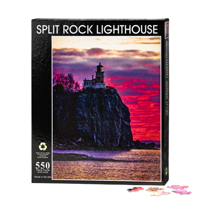 The Split Rock Sunrise by Stephanie Irwin Custom 550 piece puzzle box displays Irwin’s vibrant artwork of Split Rock Lighthouse atop a cliff at sunrise, with vivid pink, purple, and orange hues illuminating the sky above glistening water. Several loose puzzle pieces are arranged in front of the box.