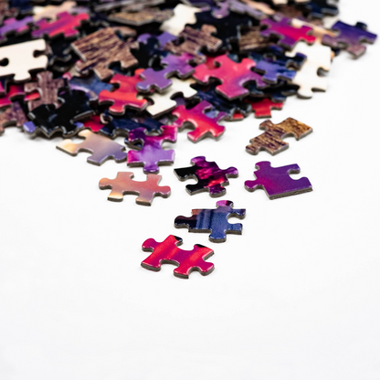 A pile of colorful jigsaw puzzle pieces from the Split Rock Sunrise by Stephanie Irwin custom 550 piece puzzle, mostly in shades of purple, red, and blue, scattered on a white surface with a few pieces separated in the foreground.