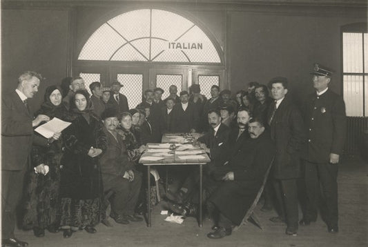 Men and women, mostly dressed in black sit and stand while filling out official forms. A police officer stands nearby. In the background a sign in the window reads "ITALIAN". Title: "Registration of Italian aliens at the Armory, St. Paul." Photo locator: E435.19 r5 in the MNHS Collections.