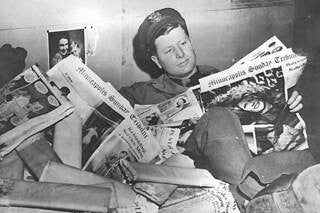 Soldier with his feet up on a desk surrounded by newspaper pages. He's reading one with interest. Title: "Minnesota soldier reading Minneapolis newspapers." 1943. Photographer: George E. Luxton. Photo locator: E448.25 p1 in the MNHS Collections.