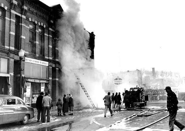 A group watches firefighters battle a smoky fire in a city building as hoses and a truck are visible. Smoke fills the scene with onlookers nearby. From MNHS collections. Order Image and Use via the MNHS Staff Image and Use Order Form.